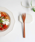 A wooden flatware fork with a warm brown finish rests on a speckled white ceramic coaster on a white surface. To the left is a white octagonal bowl filled with spaghetti, tomato sauce, and white cheese, garnished with green herbs.