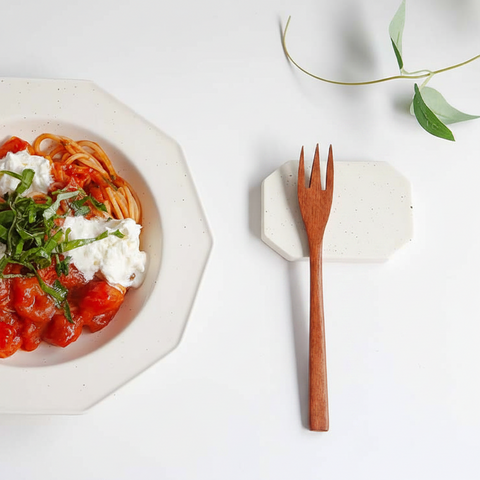 A wooden flatware fork with a warm brown finish rests on a speckled white ceramic coaster on a white surface. To the left is a white octagonal bowl filled with spaghetti, tomato sauce, and white cheese, garnished with green herbs.