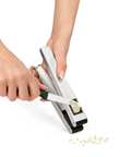 A close-up of a person's hands using a silver and black garlic press to crush garlic, with a few small garlic cloves visible on a white background.