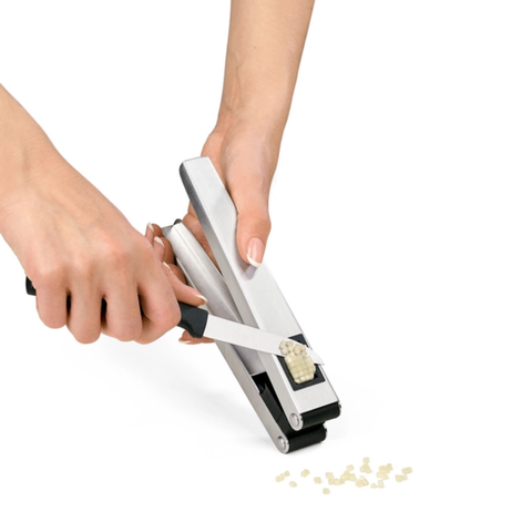 A close-up of a person's hands using a silver and black garlic press to crush garlic, with a few small garlic cloves visible on a white background.
