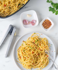 A top-down view of prepared spaghetti on a white plate with a fork, alongside a bowl of pasta, metal tongs, small dishes with garlic, red pepper flakes, and parsley on a white marble surface.