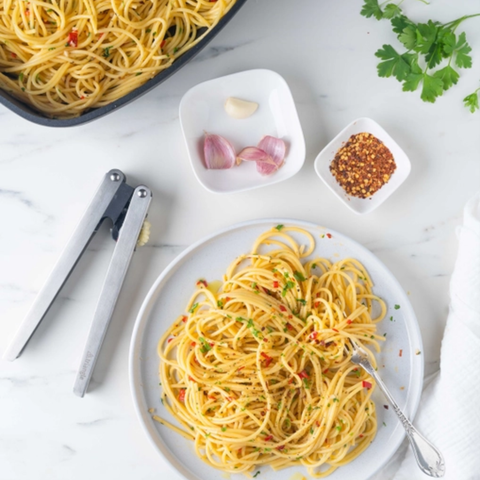 A top-down view of prepared spaghetti on a white plate with a fork, alongside a bowl of pasta, metal tongs, small dishes with garlic, red pepper flakes, and parsley on a white marble surface.