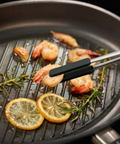 A close-up of a black grill pan with shrimp, lemon slices, rosemary, and oil on a dark background, with metal tongs holding a shrimp over the grates.