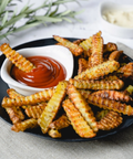Crispy golden-brown crinkle-cut fries piled on a black plate, served with a small white dish of red ketchup and a small white dish of creamy dipping sauce on a light tabletop.