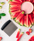 A metal watermelon slice cutter with a black handle is shown on a white surface, with sliced watermelon arranged in a circle and a small bowl of pink drink nearby. Limes, basil leaves, strawberries, and extra watermelon slices are scattered around as styling props.
