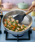 Person cooking chicken and vegetables in a pan on a stove with a spatula.