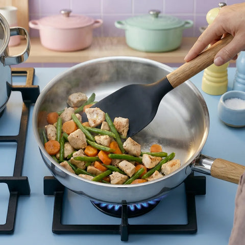 Person cooking chicken and vegetables in a pan on a stove with a spatula.