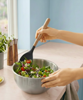 Person mixing salad with a wooden and silicone mixing spoon in a stainless steel bowl on a colorful kitchen counter