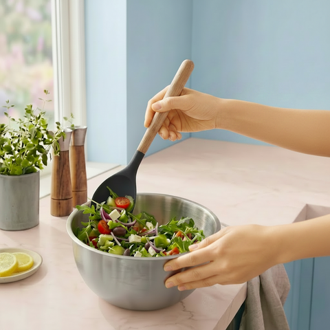 Person mixing salad with a wooden and silicone mixing spoon in a stainless steel bowl on a colorful kitchen counter