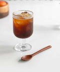 A clear stemmed glass with a thin gold rim, filled with iced coffee, sits on a white surface next to a small wooden spoon.