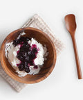 A top-down view of a wooden bowl filled with whipped cream and dark berry sauce, placed on a folded beige linen napkin with a wooden spoon on a white background.