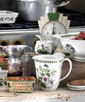 A collection of white ceramic Portmeade mugs, bowls, and jars decorated with botanical illustrations of strawberries and flowers, styled on a rustic wooden table with fresh strawberries and vintage kitchen props.