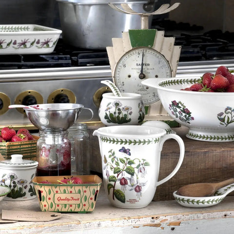 A collection of white ceramic Portmeade mugs, bowls, and jars decorated with botanical illustrations of strawberries and flowers, styled on a rustic wooden table with fresh strawberries and vintage kitchen props.