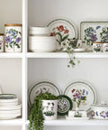 A white shelving unit displays a coordinated collection of ceramic Portmeirion Botanic Garden dishes, including plates, bowls, canisters, and a lidded box, all decorated with hand-painted purple and red flowers and green botanical borders.