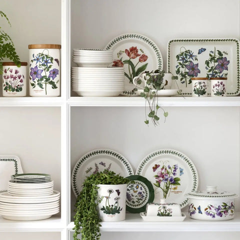 A white shelving unit displays a coordinated collection of ceramic Portmeirion Botanic Garden dishes, including plates, bowls, canisters, and a lidded box, all decorated with hand-painted purple and red flowers and green botanical borders.