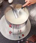 Metal cake layer tool with a hand lifting a circular metal cake layer, surrounded by baking ingredients on a dark surface.