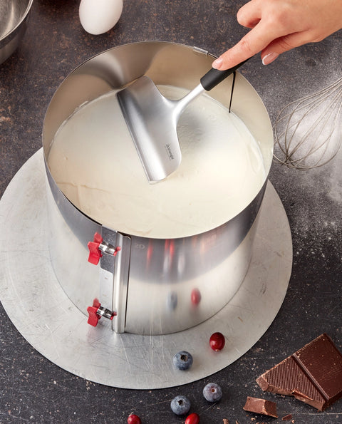 Metal cake layer tool with a hand lifting a circular metal cake layer, surrounded by baking ingredients on a dark surface.