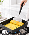 A person's hand uses a spatula to flip a rolled omelet in a black nonstick frying pan on a gas stove, with a marble kitchen countertop and green onions in a bowl in the background.