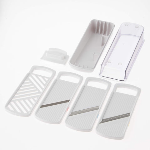 A white plastic mandoline slicer set with multiple interchangeable blade plates and a clear plastic cover, arranged on a clean white background.