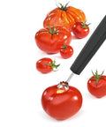 A tomato peeler and slicer tool with a black handle is shown peeling a red tomato on a white background, with additional whole tomatoes scattered behind it.