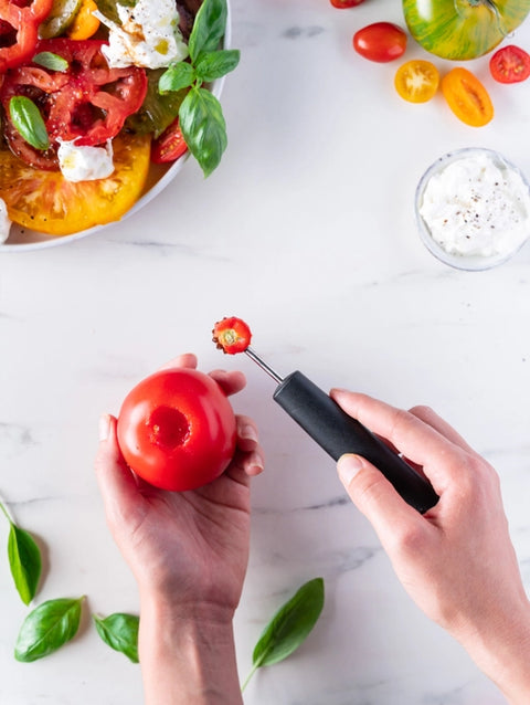 Person using a tomato corer on a red tomato with a white background featuring tomatoes and basil.