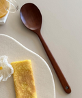 A dark brown wooden spoon with a hanging loop is displayed on a light beige surface next to a white ceramic plate with a folded omelet, a glass of orange juice, and a white flower.