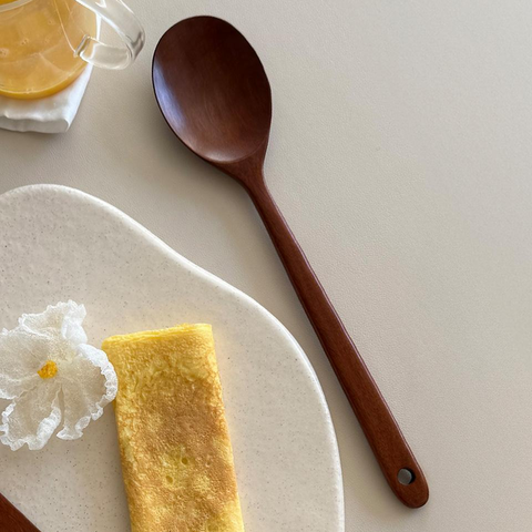 A dark brown wooden spoon with a hanging loop is displayed on a light beige surface next to a white ceramic plate with a folded omelet, a glass of orange juice, and a white flower.
