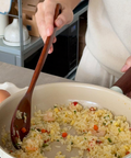 A person cooking fried rice in a beige non-stick pan on a stovetop, using a wooden spoon. The kitchen background includes a shelf with kitchen items and a carton.