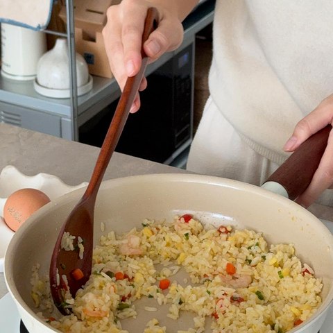 A person cooking fried rice in a beige non-stick pan on a stovetop, using a wooden spoon. The kitchen background includes a shelf with kitchen items and a carton.