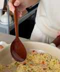 A person in a cream sweater uses a wooden spoon to stir-fry rice in a beige pan in a kitchen setting.
