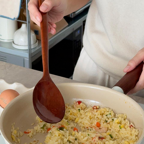 A person in a cream sweater uses a wooden spoon to stir-fry rice in a beige pan in a kitchen setting.