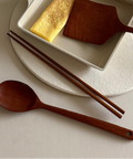 A beige square frying pan with a white handle contains a folded omelet and a wooden spatula, styled on a matching beige plate with wooden chopsticks and a wooden spoon on a light neutral background.