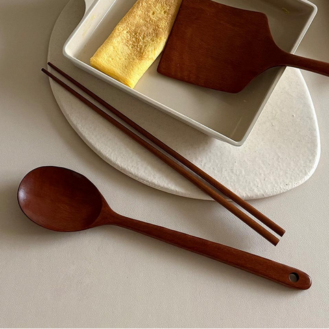 A beige square frying pan with a white handle contains a folded omelet and a wooden spatula, styled on a matching beige plate with wooden chopsticks and a wooden spoon on a light neutral background.