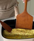 A beige squarel pan on a white electric burner with an omelette cooking inside, alongside two wooden spatulas. A person wearing a beige apron is visible at the top of the frame.