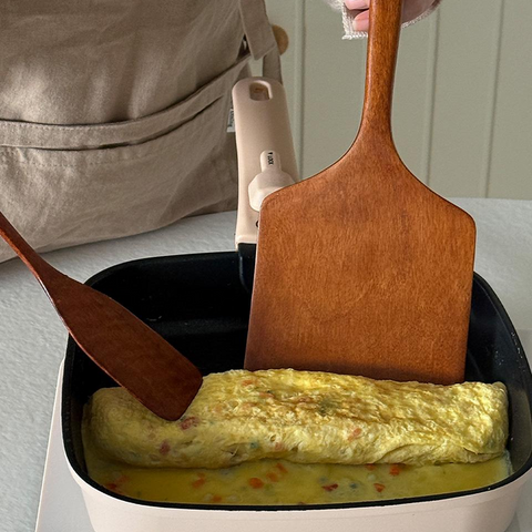 A beige squarel pan on a white electric burner with an omelette cooking inside, alongside two wooden spatulas. A person wearing a beige apron is visible at the top of the frame.