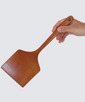 A wooden pizza peel with a warm brown finish, held by a person's hand against a solid light gray background.