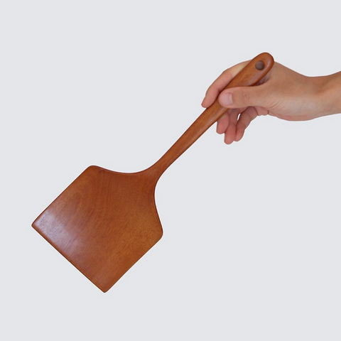 A wooden pizza peel with a warm brown finish, held by a person's hand against a solid light gray background.