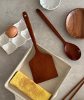 A wooden spatula and wooden spoon set arranged on a gray stone surface, with a folded yellow omelet in a beige rectangular dish and a brown egg in a white egg carton nearby.