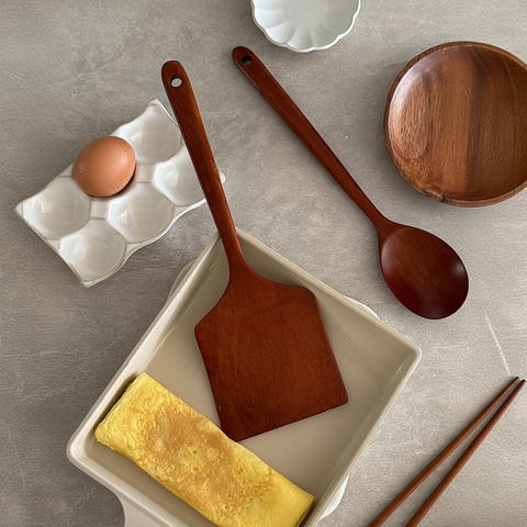 A wooden spatula and wooden spoon set arranged on a gray stone surface, with a folded yellow omelet in a beige rectangular dish and a brown egg in a white egg carton nearby.