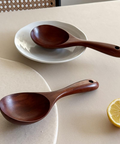 Two dark brown wooden serving spoons with hanging holes, displayed on a beige table with a white plate, a lemon slice, and a woven chair back in the background.