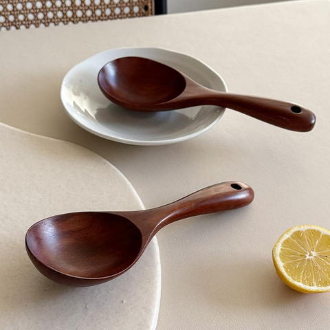 Two dark brown wooden serving spoons with hanging holes, displayed on a beige table with a white plate, a lemon slice, and a woven chair back in the background.