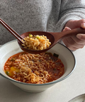 A person in a gray knit sweater holds a wooden spoon filled with cooked ramen noodles above a bowl of spicy red instant ramen. Chopsticks rest on the bowl, and a plate of sushi is partially visible at the lower right on a light tabletop.