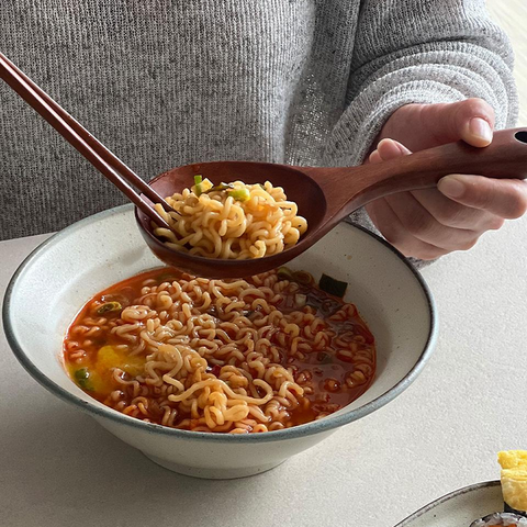 A person in a gray knit sweater holds a wooden spoon filled with cooked ramen noodles above a bowl of spicy red instant ramen. Chopsticks rest on the bowl, and a plate of sushi is partially visible at the lower right on a light tabletop.