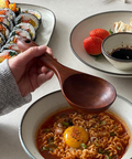 A hand holds a wooden spoon over a bowl of spicy ramen with a raw egg yolk, with plates of sushi and dumplings in the background on a light tabletop.