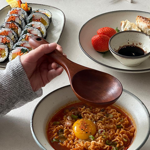 A hand holds a wooden spoon over a bowl of spicy ramen with a raw egg yolk, with plates of sushi and dumplings in the background on a light tabletop.