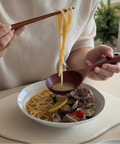 A close-up of a person using wooden chopsticks and a spoon to serve pasta and clams from a white bowl on a light beige table. The person is wearing a cream-colored knit sweater, with a blurred green plant in the background.