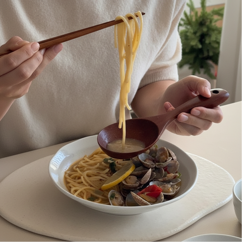 A close-up of a person using wooden chopsticks and a spoon to serve pasta and clams from a white bowl on a light beige table. The person is wearing a cream-colored knit sweater, with a blurred green plant in the background.