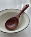 A dark reddish-brown wooden serving spoon with a hole in the handle, resting inside a speckled beige ceramic bowl on a white surface.