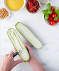 A top-down view of a person using a vegetable peeler to remove the skin from a halved zucchini on a white marble countertop, surrounded by bowls of ingredients including cherry tomatoes, basil, sun-dried tomatoes, and grains.