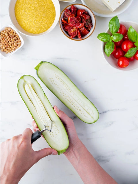 A top-down view of a person using a vegetable peeler to remove the skin from a halved zucchini on a white marble countertop, surrounded by bowls of ingredients including cherry tomatoes, basil, sun-dried tomatoes, and grains.